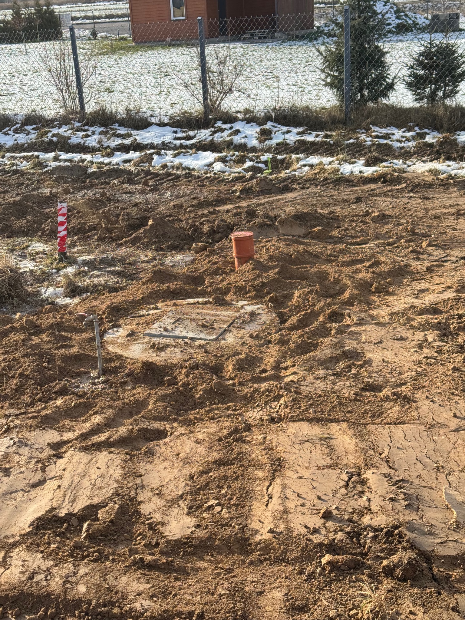 Teren po robotach ziemnych z widocznym włazem kanalizacyjnym, rurą PCV i palikiem oznaczającym przebieg instalacji, w tle ogrodzenie i częściowo ośnieżony teren.