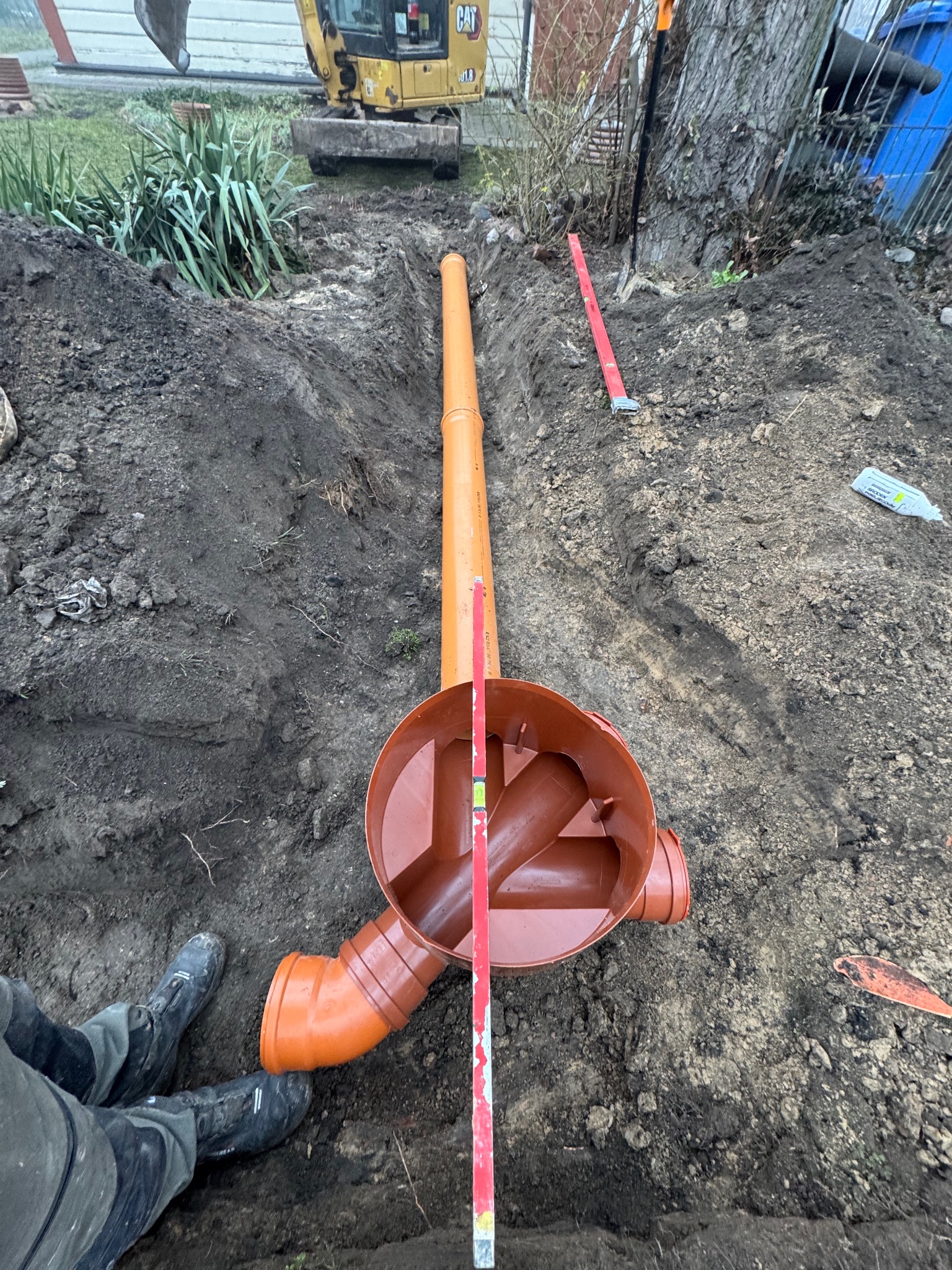 Pomarańczowe rury kanalizacyjne z kształtkami ułożone w wykopie, poziomica oparta o kształtkę, w tle minikoparka CAT, ziemia, trawa, krzewy, płot i drzewo.