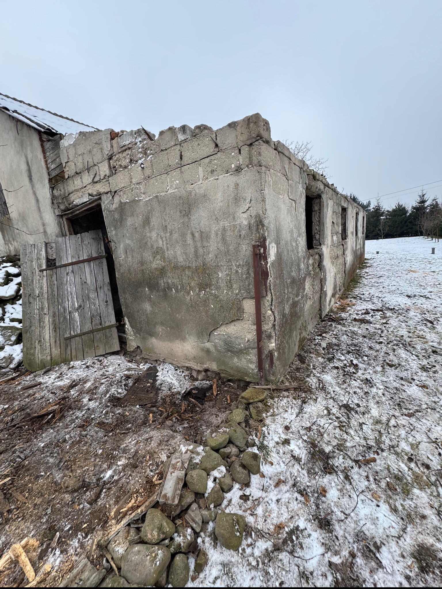 Zdewastowany, częściowo rozebrany budynek z betonu i cegieł, z otwartymi drewnianymi drzwiami, otoczony śniegiem i gruzem, w tle widoczne drzewa.