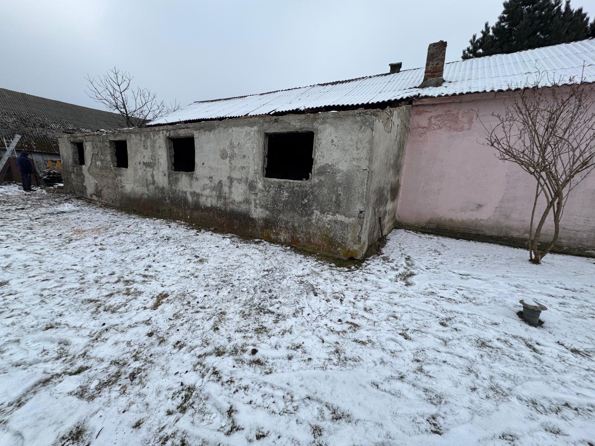 Niedokończony budynek z otworami okiennymi bez szyb, pokryty śniegiem, z fragmentem różowej ściany i widocznym kominem.