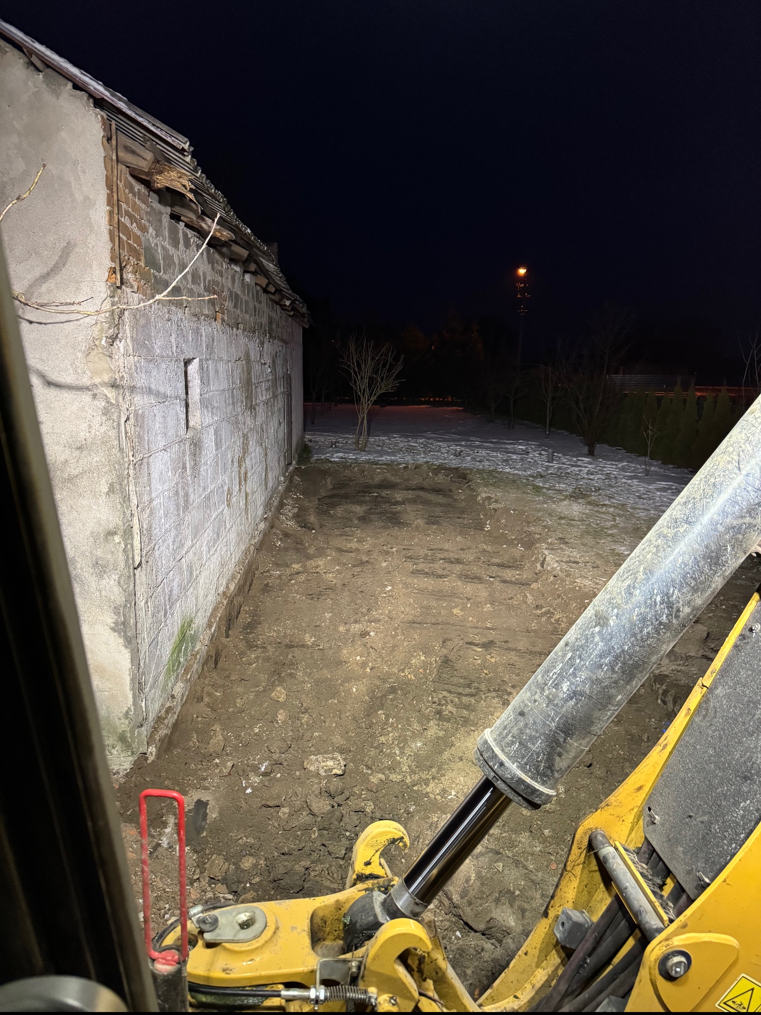 Wykop pod fundamenty starego budynku z pustaków, widok z kabiny żółtej koparki w nocy, oświetlony teren budowy.