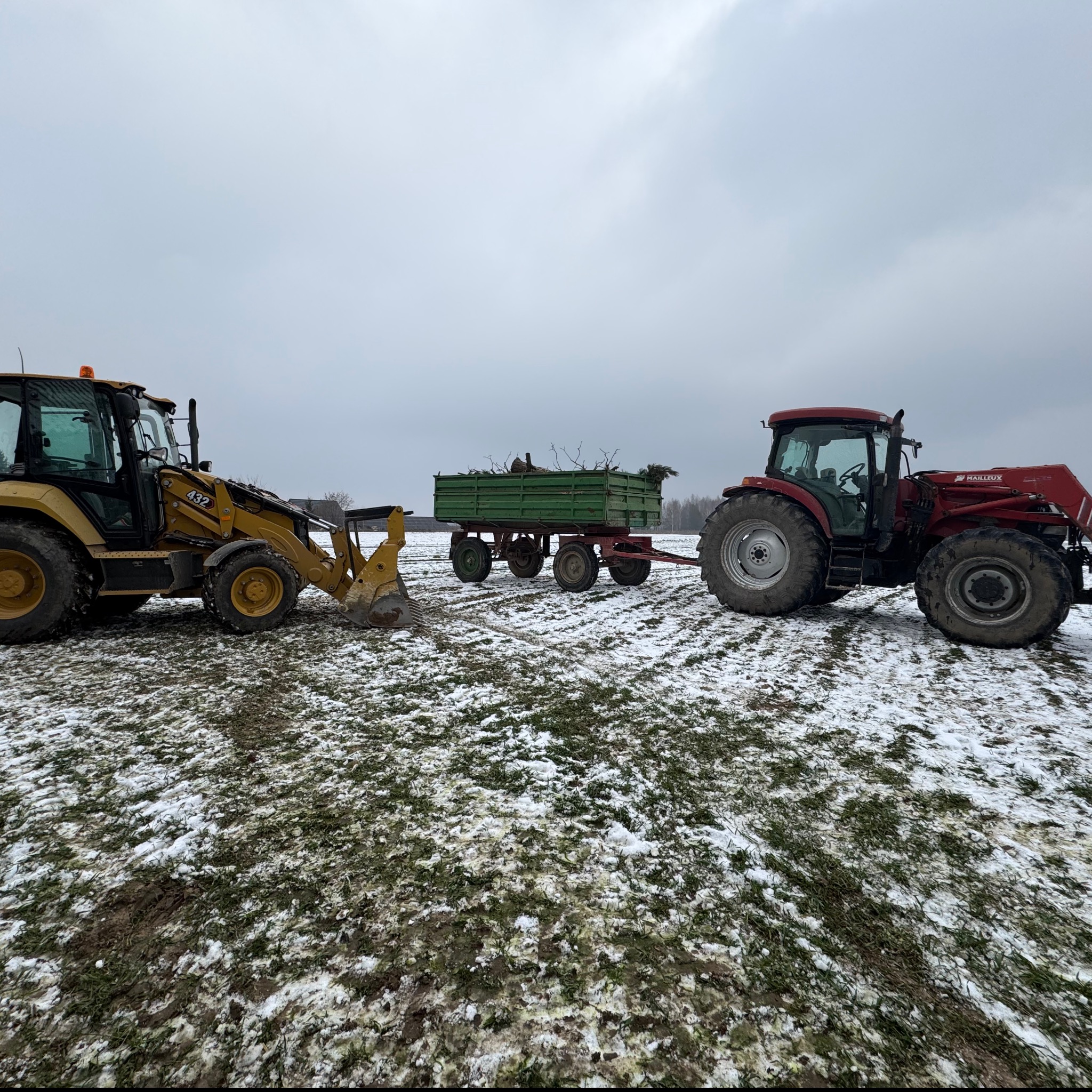 Żółta ładowarka i czerwony traktor z przyczepą wypełnioną gałęziami na zasypanym śniegiem polu w pochmurny dzień.