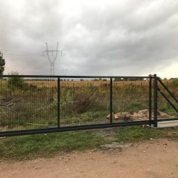FENTA Ogrodzenia - Przesuwna brama wjazdowa z czarną metalową ramą i wypełnieniem z siatki, zamontowana na betonowych słupkach, widok na polną okolicę i słupy wysokiego napięcia pod pochmurnym niebem.