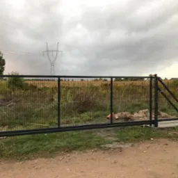 Przesuwna brama wjazdowa z czarną metalową ramą i wypełnieniem z siatki, zamontowana na betonowych słupkach, widok na polną okolicę i słupy wysokiego napięcia pod pochmurnym niebem.