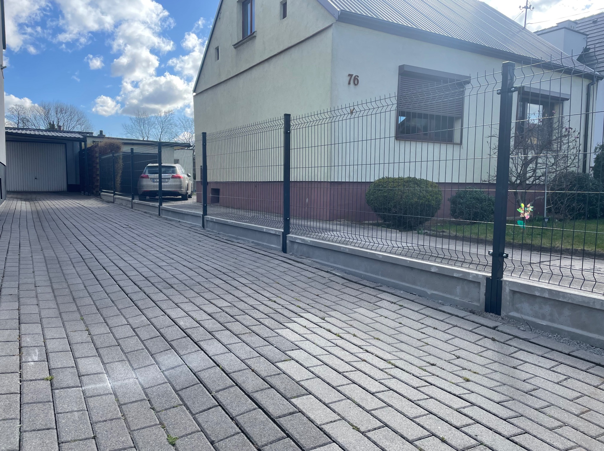 Nowoczesne, czarne ogrodzenie panelowe z podmurówką betonową, widok z perspektywy chodnika z kostki brukowej, w tle budynek mieszkalny z numerem 76 i zaparkowany samochód.