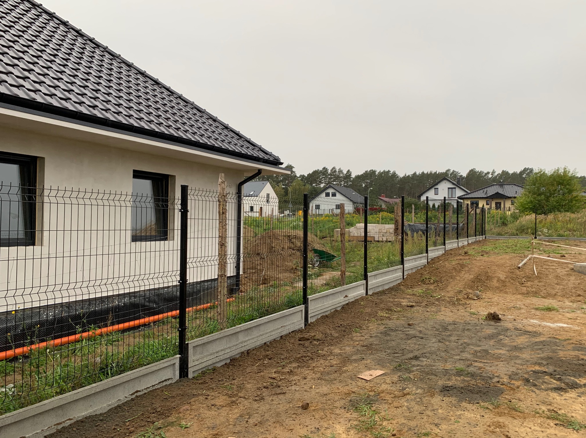 Nowoczesne ogrodzenie z paneli z czarnymi słupkami i betonową podmurówką, widok z boku domu z czarnymi oknami, w tle inne domy i drzewa.