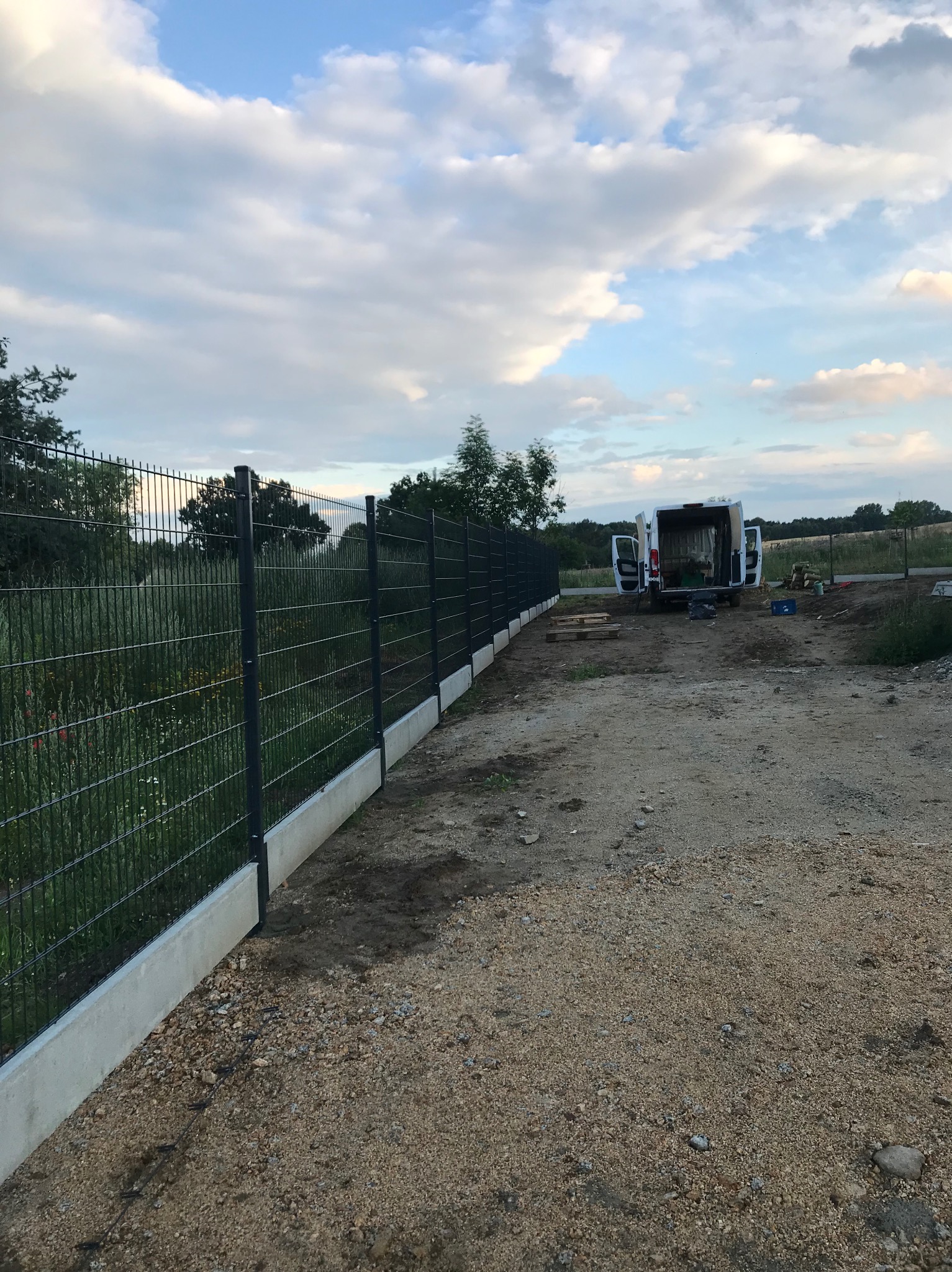 Długi, ciemny płot z siatki panelowej na betonowej podmurówce, widoczny z boku, z otwartym busem w tle i narzędziami budowlanymi, na ziemnej nawierzchni w słoneczny dzień.