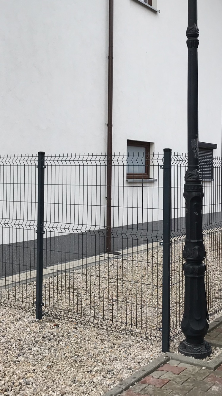 Nowoczesne, grafitowe ogrodzenie z paneli zgrzewanych, oddzielające posesję od chodnika z kostki, widok na białą ścianę budynku z oknem i starą, czarną latarnię uliczną.