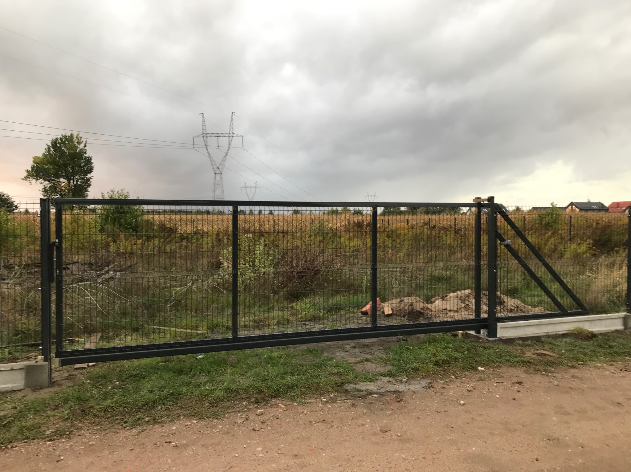 Przesuwna brama wjazdowa z czarną metalową ramą i wypełnieniem z siatki, zamontowana na betonowych słupkach, widok na polną okolicę i słupy wysokiego napięcia pod pochmurnym niebem.
