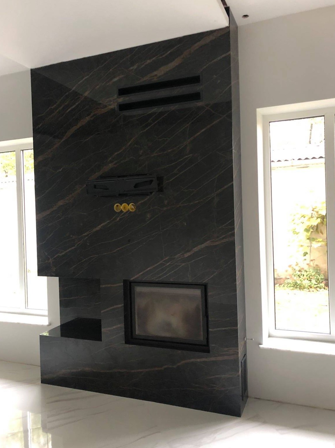 Wykończony kominek z czarnego marmuru z złotymi żyłkami, z wbudowanym wkładem kominkowym i kratkami wentylacyjnymi, w jasnym wnętrzu z białymi oknami.