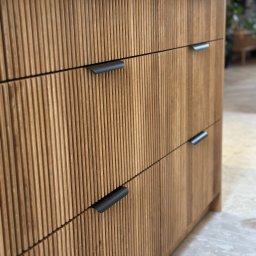 Och Meble Olaf Chmura - Szczegółowe ujęcie frontu szuflad z ryflowanego drewna w odcieniach beżu i brązu, z minimalistycznymi czarnymi uchwytami, na tle jasnej podłogi i rozmytego wnętrza.