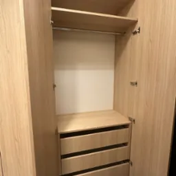 Otwarta szafa z jasnego drewna z trzema szufladami, p&oacute;łkami i drążkiem na wieszaki, widoczne okucia i zawiasy.