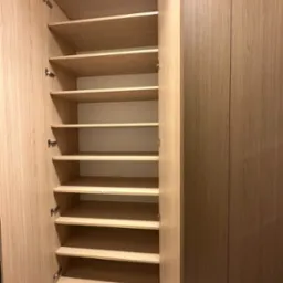 Otwarta szafa wnękowa z jasnego drewna z wieloma półkami, widoczne zawiasy.