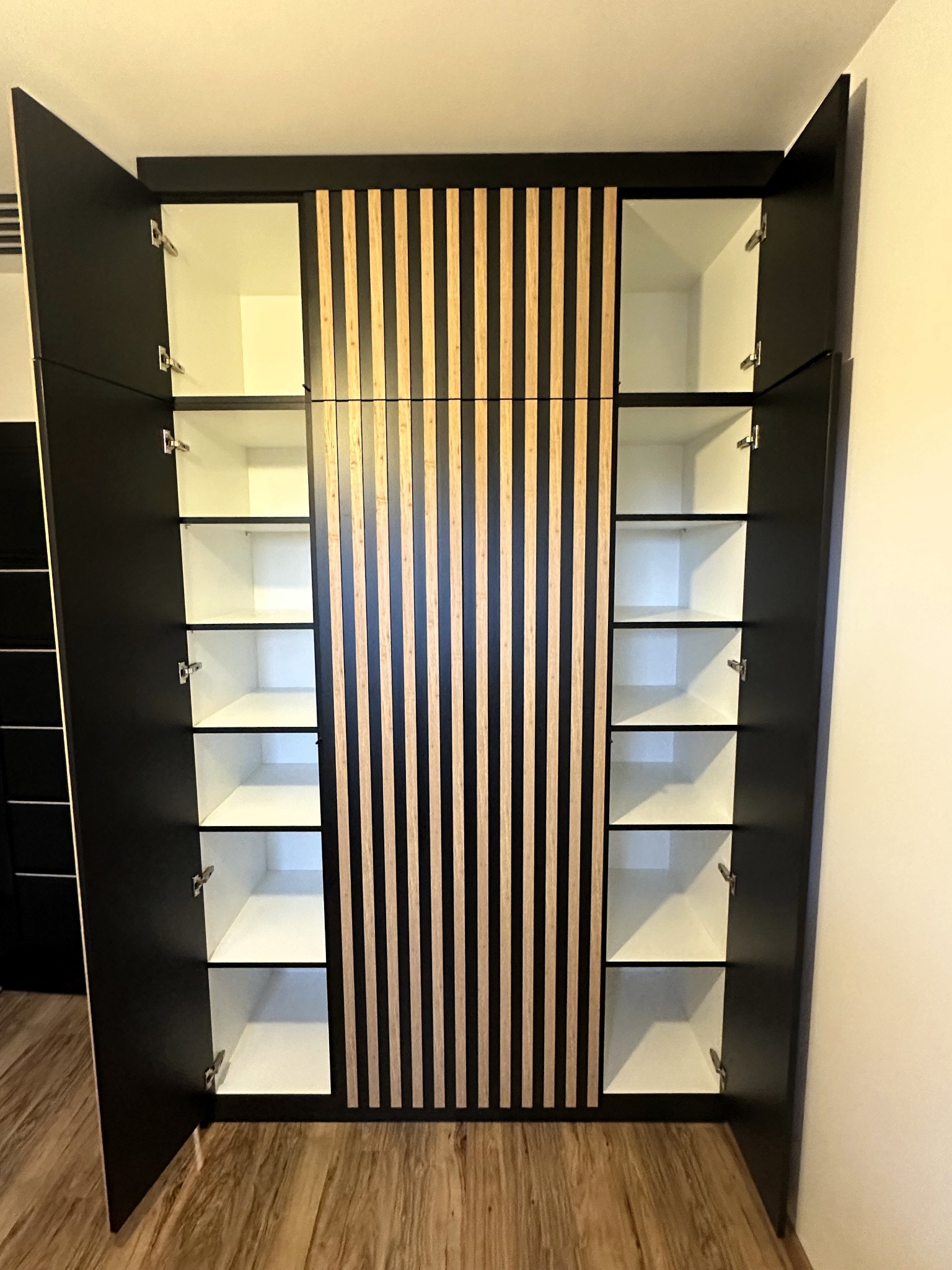 Czarna szafa wnękowa z drewnianymi lamelami pośrodku i białymi półkami wewnątrz, z otwartymi drzwiami, zrealizowana we Wrocławiu.