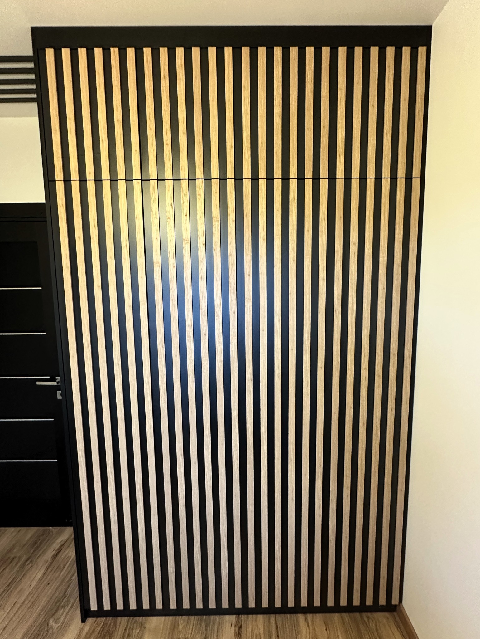 Nowoczesna szafa wnękowa z frontami z pionowych, drewnianych listew w kolorze jasnego drewna na czarnym tle, obok widoczny fragment czarnych drzwi.