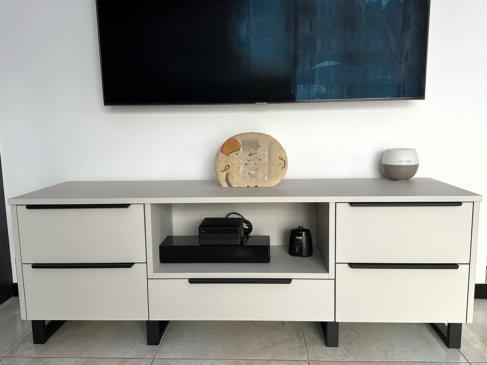 Biała szafka RTV z szufladami i otwartą półką, czarne podłużne uchwyty, na półce dekoder i nawilżacz powietrza, nad szafką wisi czarny telewizor.