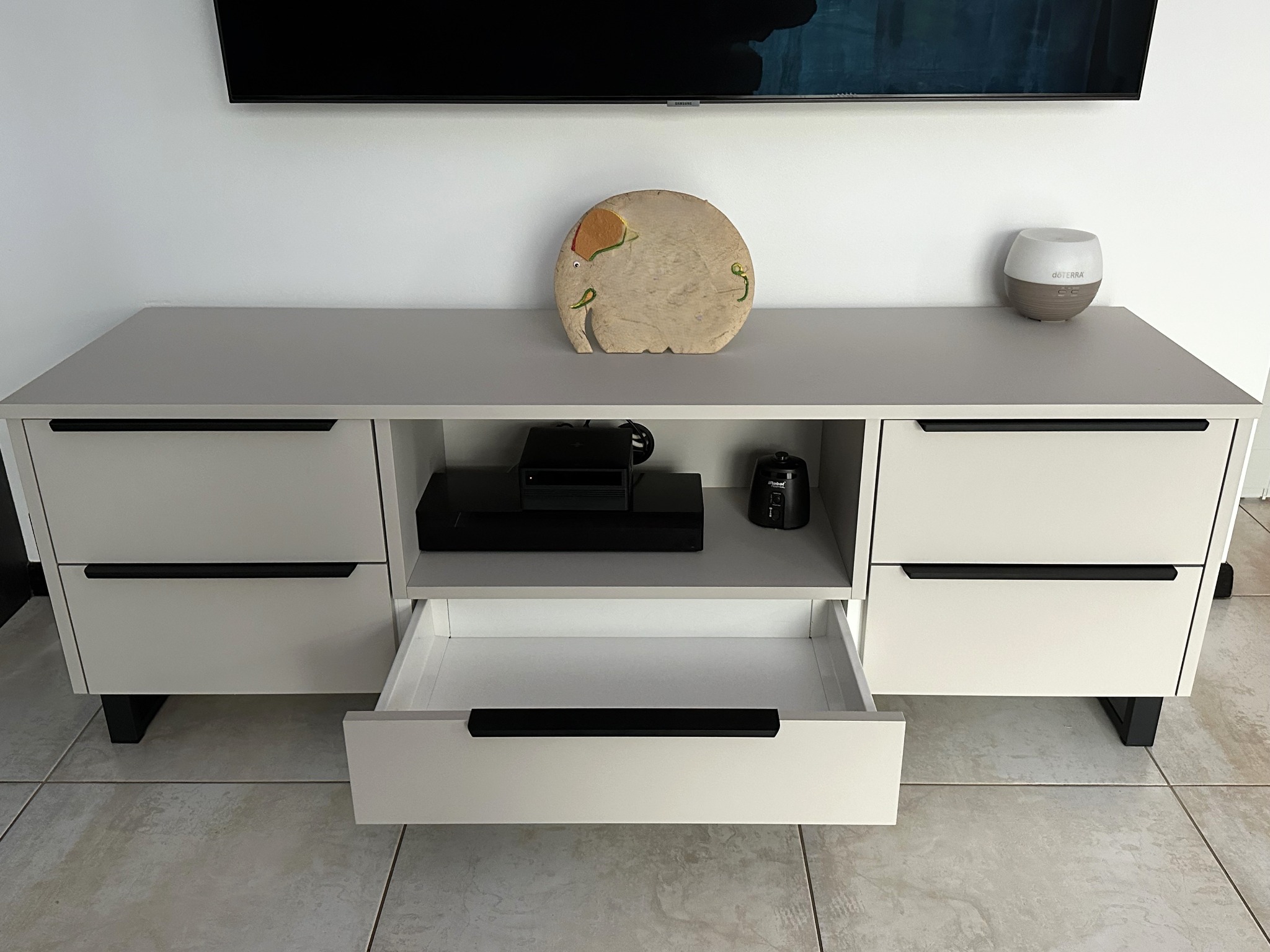 Biała szafka RTV z otwartą szufladą, czarnymi uchwytami i nóżkami, na blacie drewniany słonik i dyfuzor zapachowy, nad szafką zawieszony telewizor.