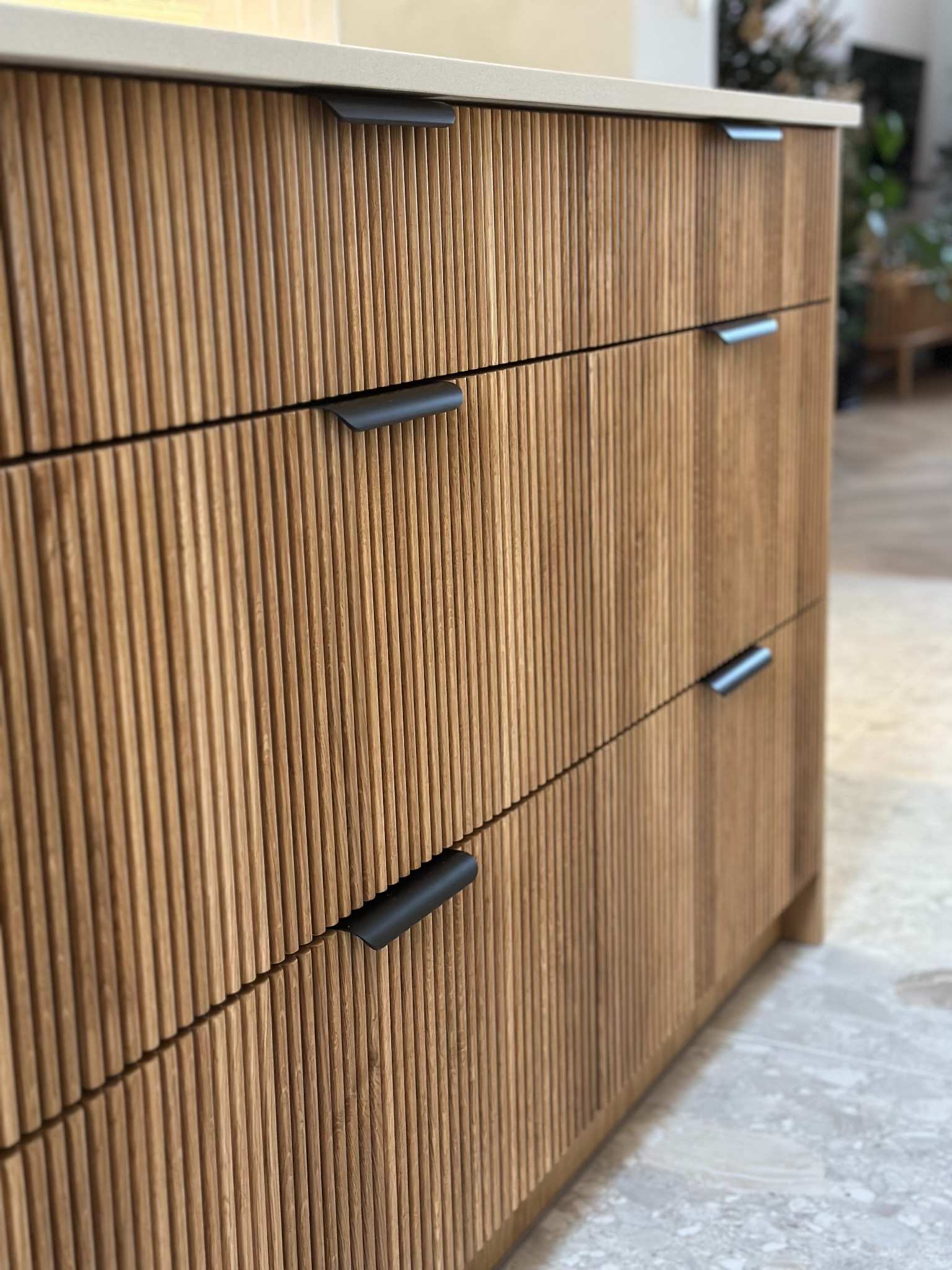 Szczegółowe ujęcie frontu szuflad z ryflowanego drewna w odcieniach beżu i brązu, z minimalistycznymi czarnymi uchwytami, na tle jasnej podłogi i rozmytego wnętrza.