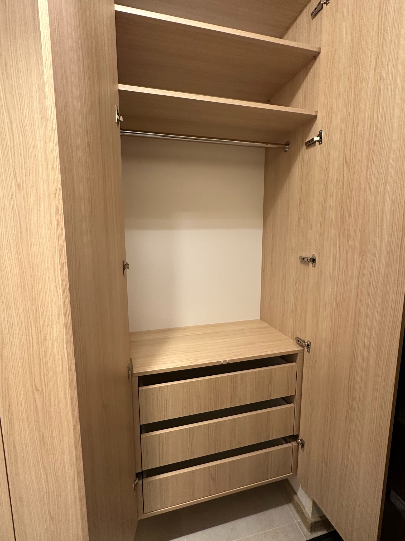 Otwarta szafa z jasnego drewna z trzema szufladami, półkami i drążkiem na wieszaki, widoczne okucia i zawiasy.