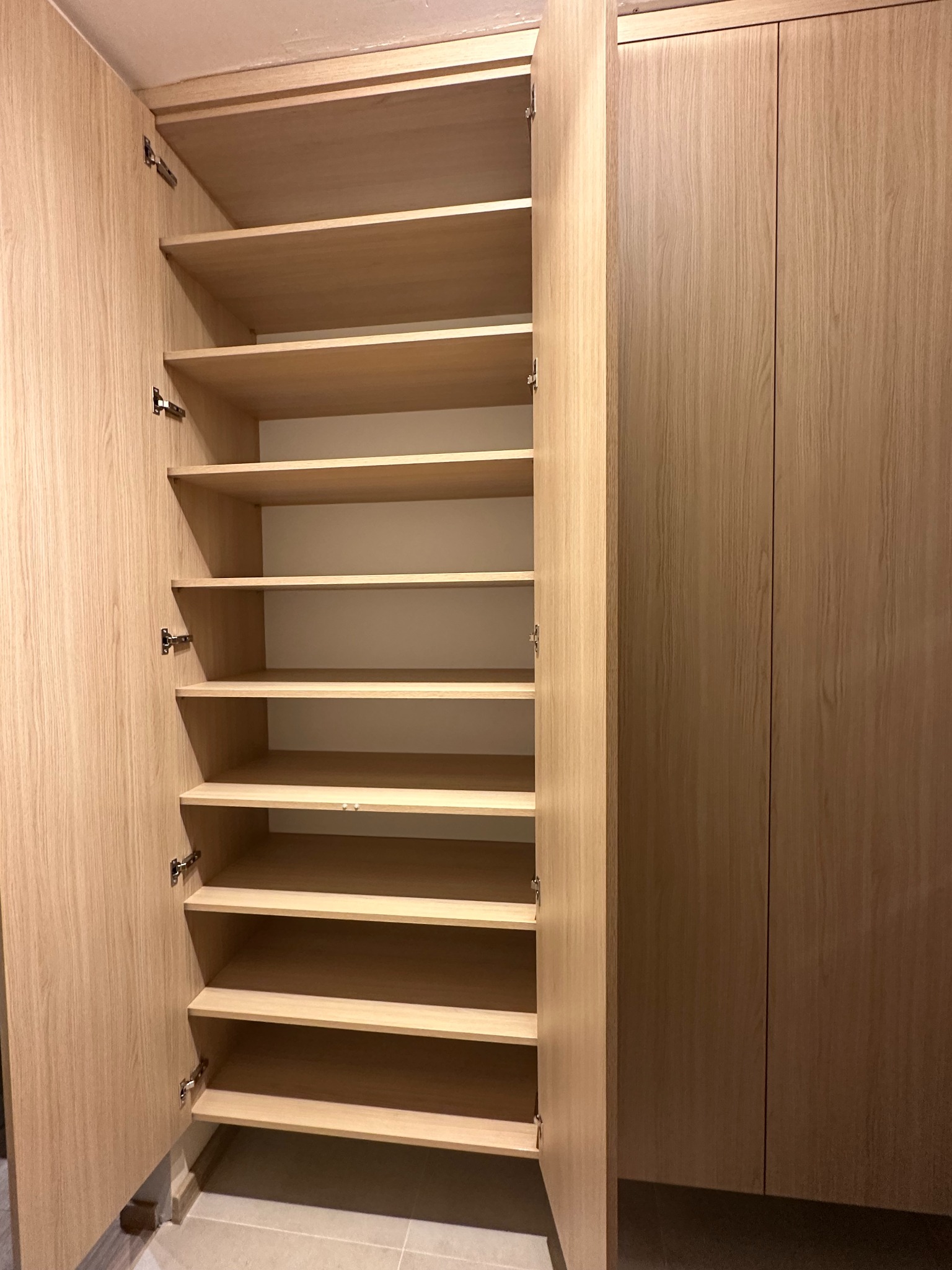 Otwarta szafa wnękowa z jasnego drewna z wieloma półkami, widoczne zawiasy.