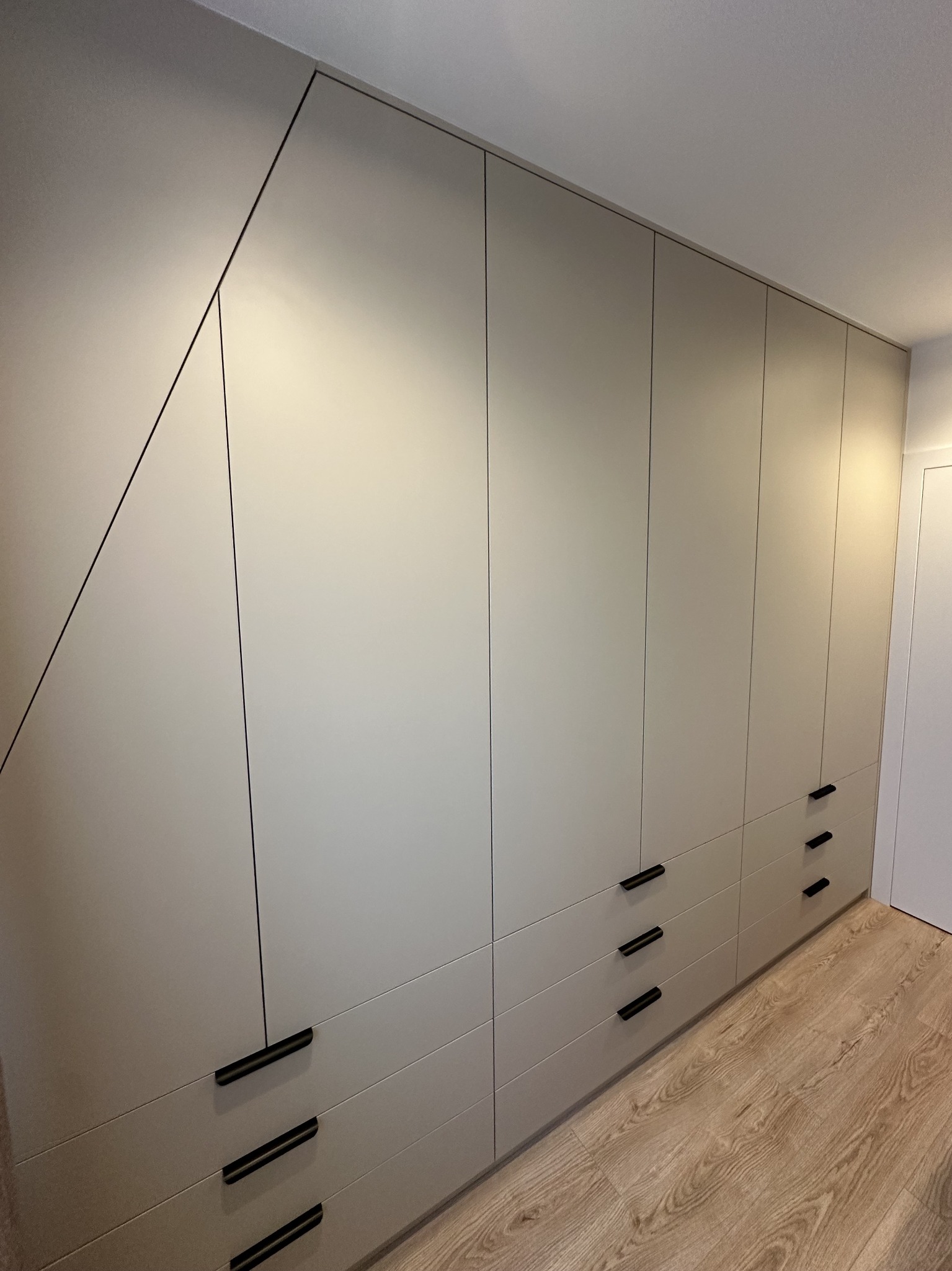 Jasnoszara szafa wnękowa z czarnymi uchwytami, dopasowana do skosu sufitu, z widoczną podłogą w odcieniach drewna.