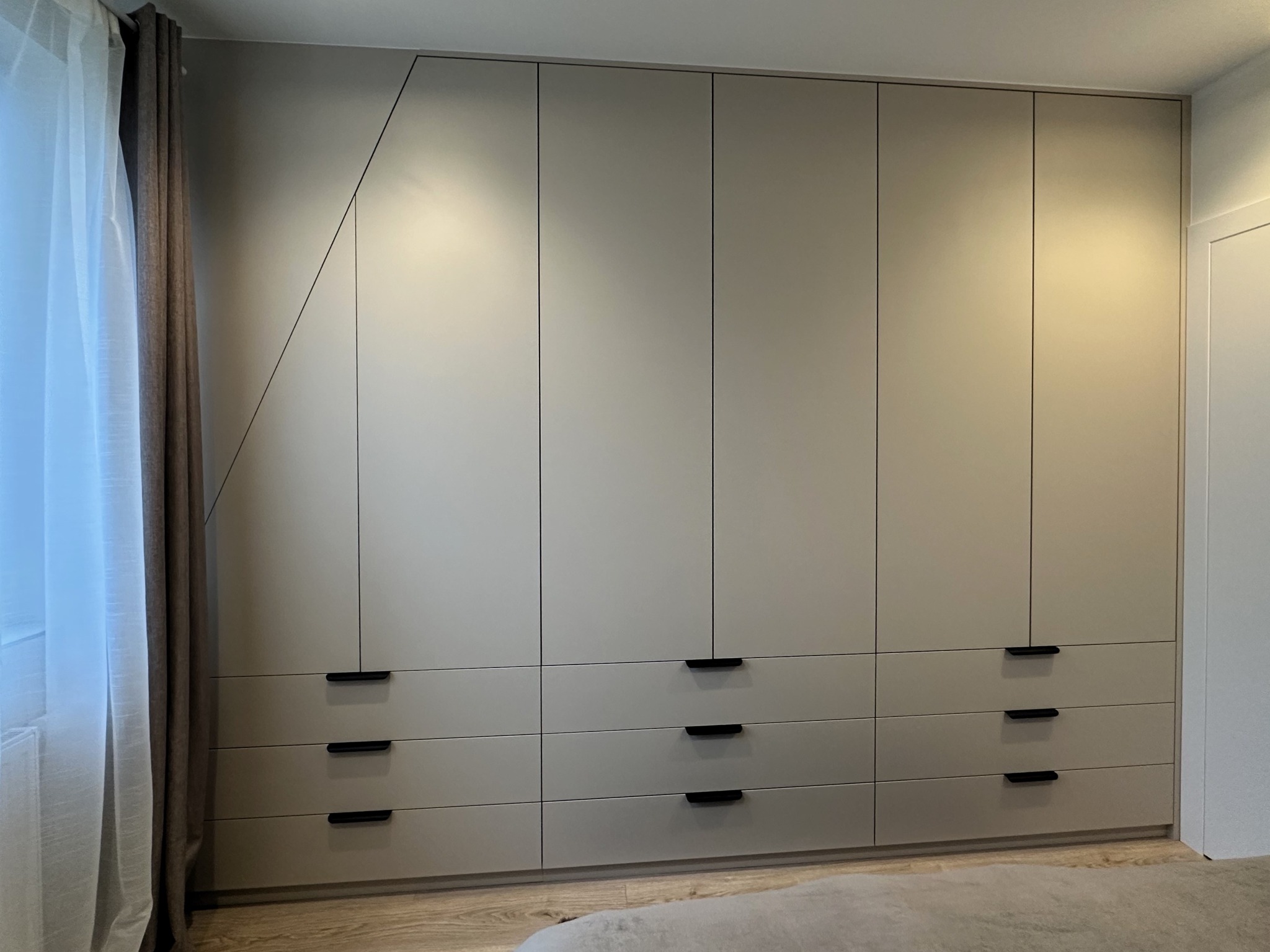 Beżowa szafa wnękowa z czarnymi uchwytami, z asymetrycznym podziałem frontów, widoczna z perspektywy podłogi, z zasłoniętym oknem po lewej stronie.