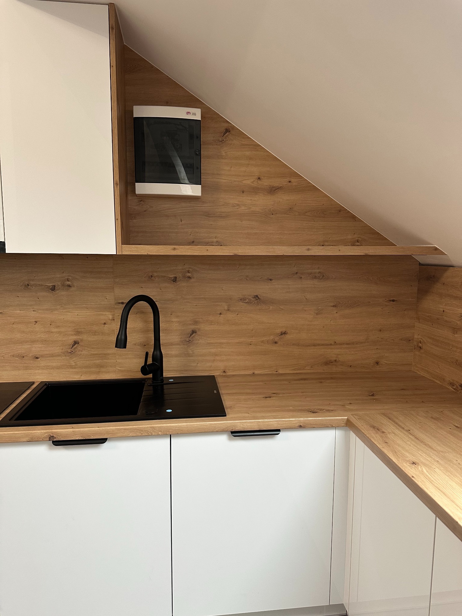 Nowoczesna kuchnia z białymi szafkami i blatem imitującym drewno, czarny zlew i bateria, zabudowa pod skosem dachu.