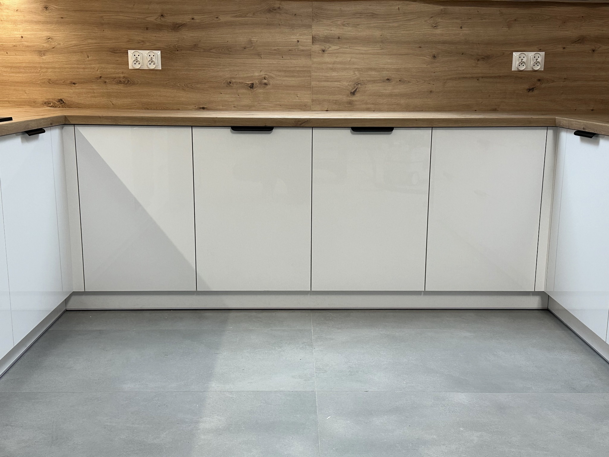 Minimalistyczna zabudowa kuchenna w kształcie litery U z białymi, lakierowanymi frontami szafek i drewnianym blatem z widocznymi gniazdkami elektrycznymi, szara podłoga z płytek.