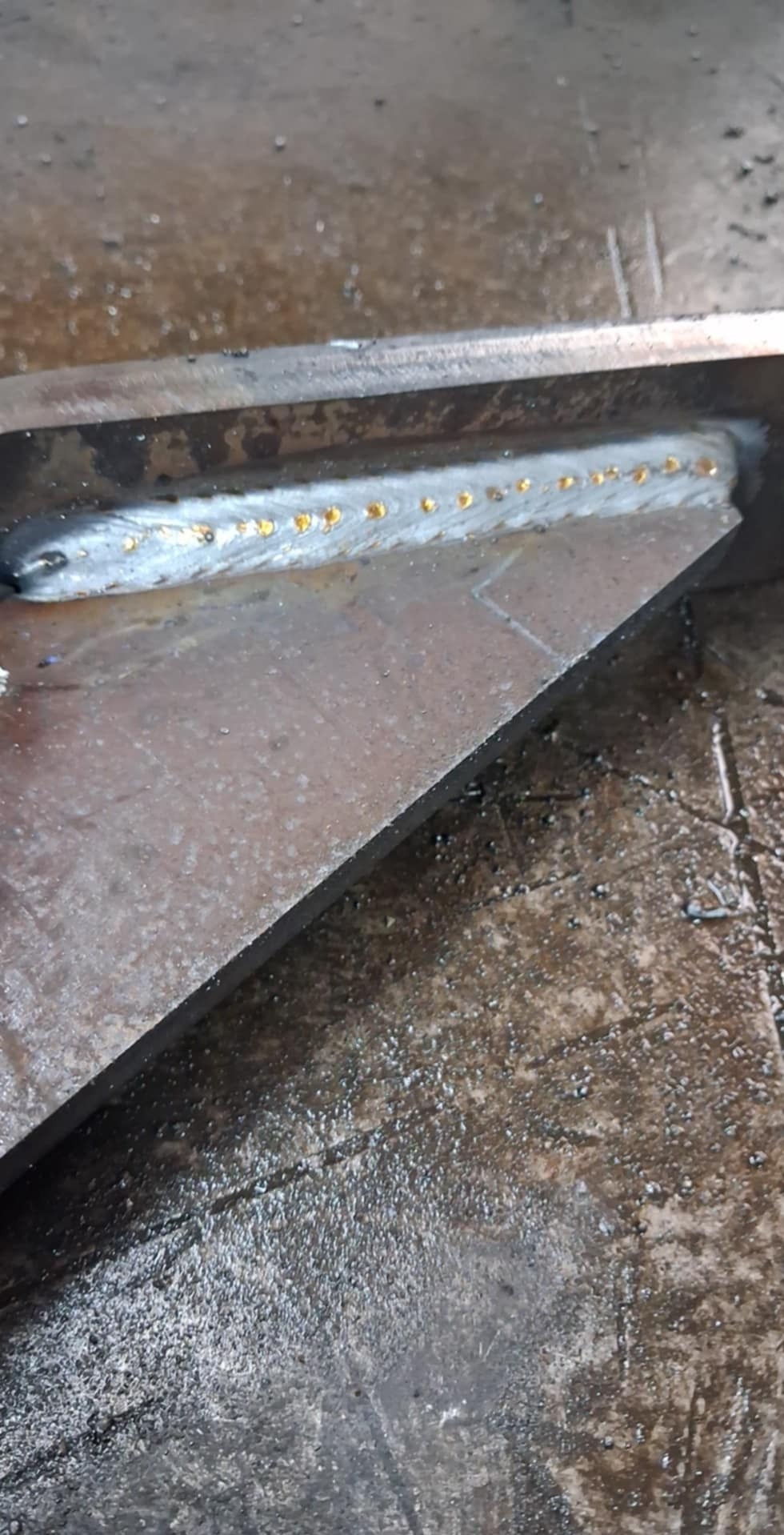 Spoina na metalowym profilu kątowym, widoczne złote krople stopionego metalu wzdłuż linii spawu, surowy, przemysłowy wygląd.