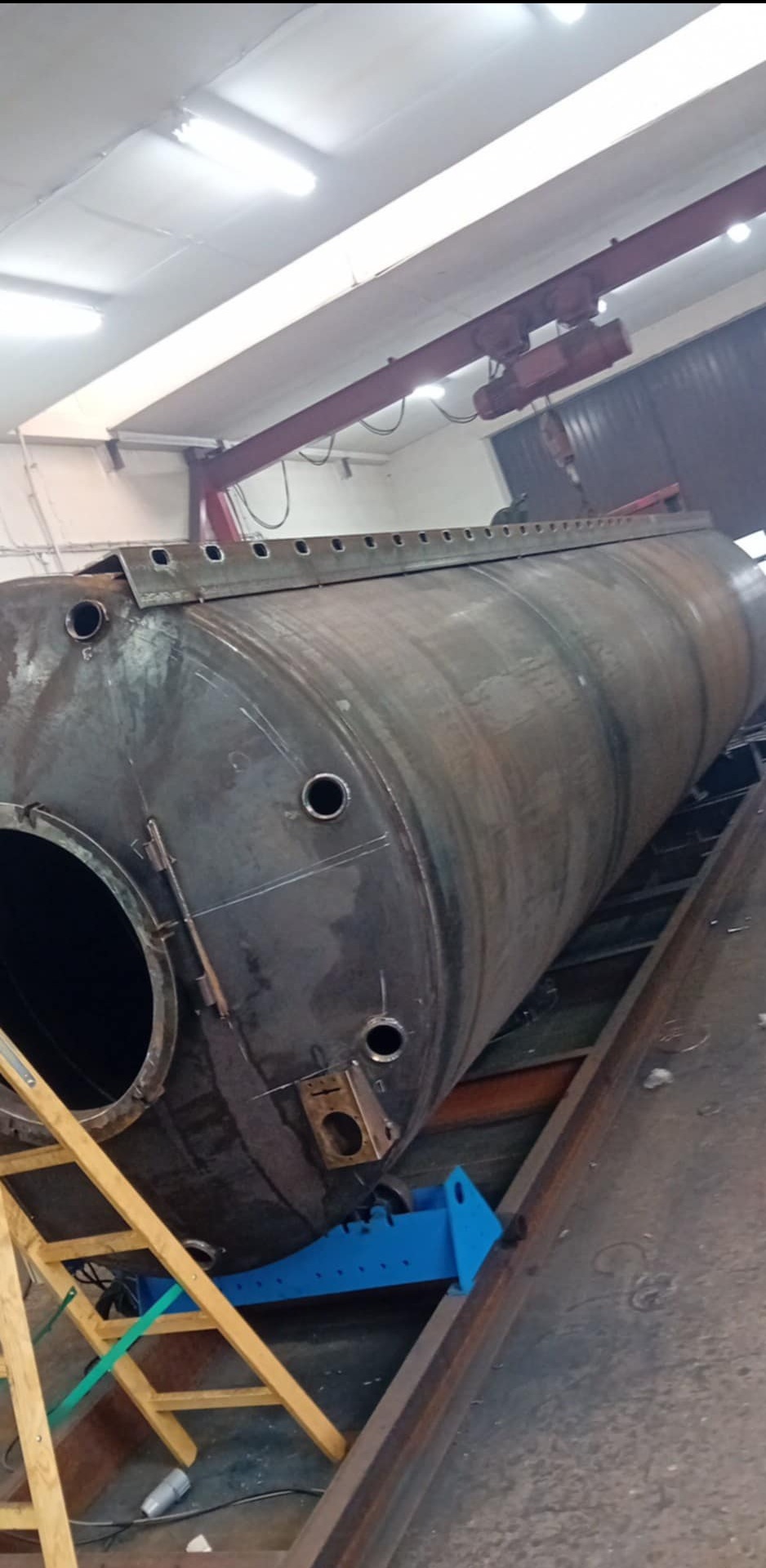 Duży, metalowy zbiornik cylindryczny z otworami i elementami mocującymi, umieszczony na konstrukcji wsporczej w hali produkcyjnej. Widoczny żuraw suwnicowy pod sufitem.