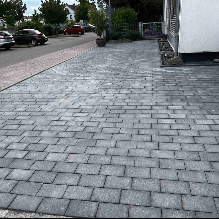 Ułożona szara kostka brukowa tworząca podjazd, z widocznymi drzewami i samochodami w tle, oraz fragmentem budynku po prawej stronie.