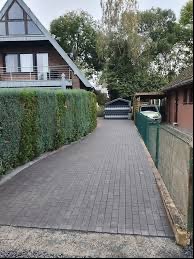 Szara kostka brukowa ułożona na podjeździe do domu jednorodzinnego, widoczny żywopłot po lewej stronie i ogrodzenie z siatki po prawej, w tle garaż i samochód.