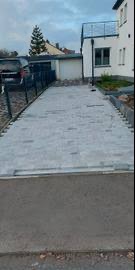 Ułożona szara kostka brukowa na podjeździe przed domem z jasną elewacją i czarną balustradą balkonu, widok z dołu w pochmurny dzień.