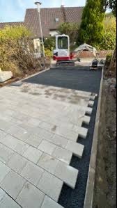 Fragment nowej nawierzchni z jasnoszarej kostki brukowej ułożonej we wzór jodełki, częściowo podsypanej ciemnym grysem, z minikoparką w tle i widocznym fragmentem ogrodu.