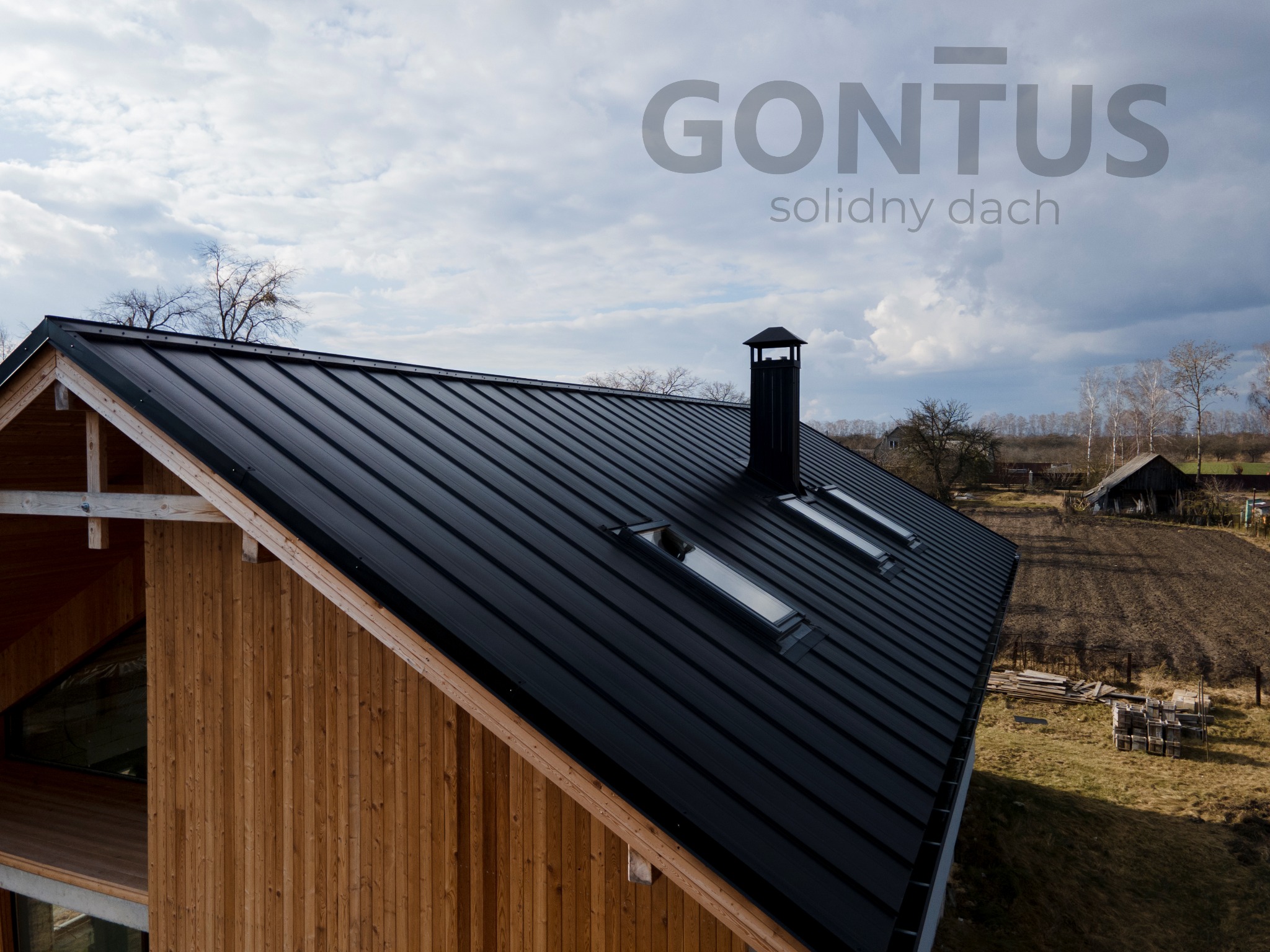 Czarny, blaszany dach z trzema oknami dachowymi i kominem na drewnianym domu z widocznym logo firmy 'GONTUS' na pochmurnym niebie.