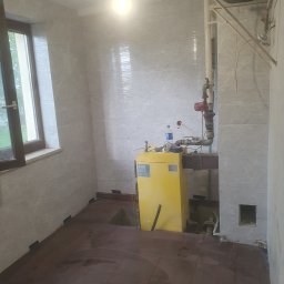 Usługi Remontowo Budowlane - Wnętrze łazienki w trakcie prac remontowych, widoczne płytki imitujące marmur na ścianach, ciemne płytki podłogowe z wyciętymi fragmentami, żółty piec grzewczy oraz instalacja hydrauliczna...