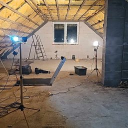 Usługi Remontowo Budowlane - Poddasze w trakcie adaptacji: ocieplenie wełną mineralną między krokwiami, surowe ściany z bloczków, drabina, reflektory budowlane i prowizoryczne schody z desek.
