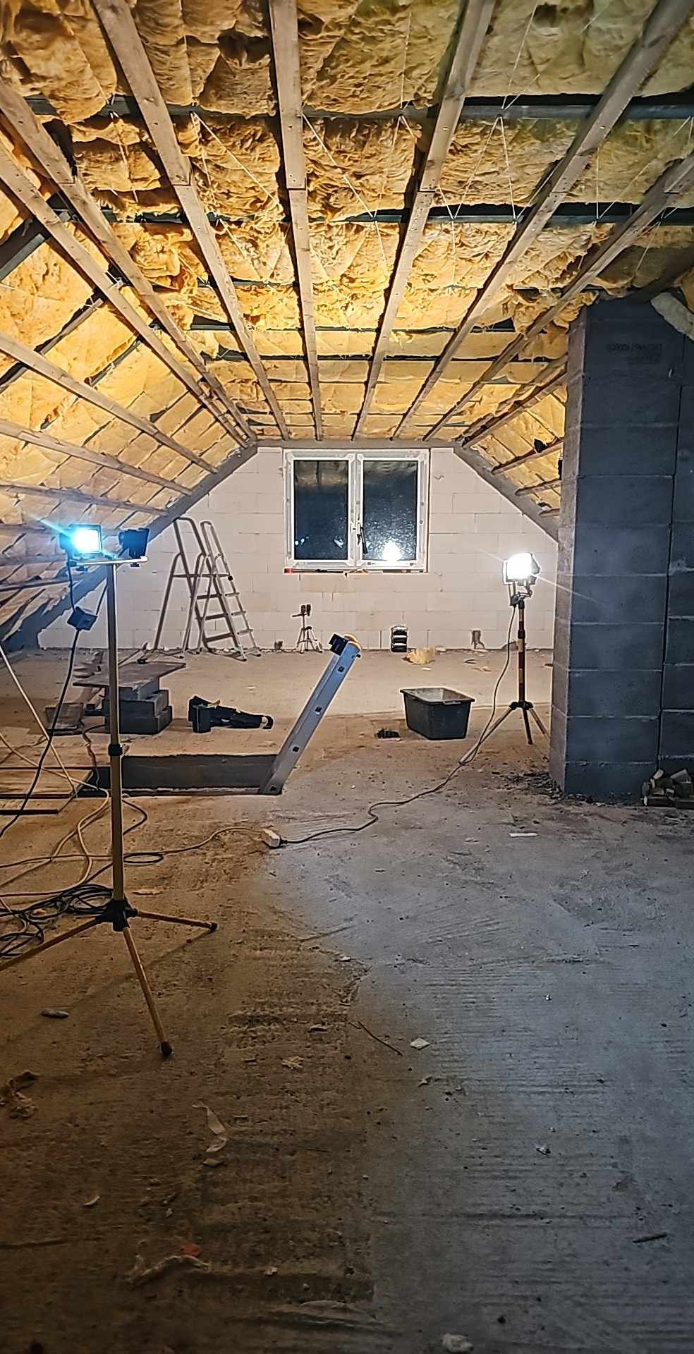 Poddasze w trakcie adaptacji: ocieplenie wełną mineralną między krokwiami, surowe ściany z bloczków, drabina, reflektory budowlane i prowizoryczne schody z desek.