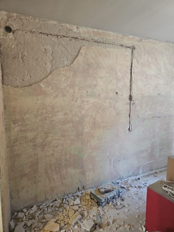 Ściana w trakcie przygotowania do prac wykończeniowych, z widocznymi bruzdami pod instalacje elektryczne i częściowo usuniętą starą warstwą tynku, gruz na podłodze i skrzynka z narzędziami.