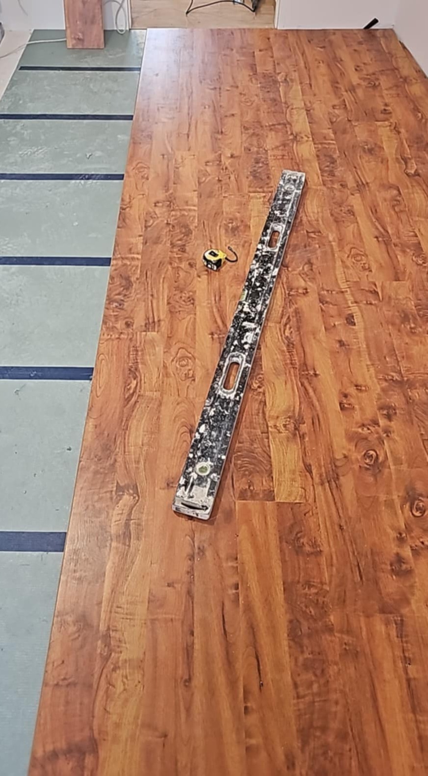 Ułożone panele podłogowe o wzorze drewna, poziomica z zabrudzeniami oraz zwinięta miarka leżą na podłodze, widoczny fragment podkładu pod panele.