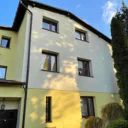 Dwukondygnacyjny dom z elewacją w kolorze żółtym i kremowym, z ciemnymi oknami i rynnami, otoczony zielenią, z widocznymi śladami cieni drzew na ścianie.