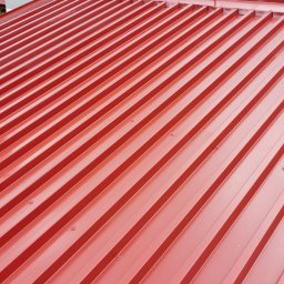 MDE Malowanie Dachów i Elewacji - Czerwony dach z blachy trapezowej po renowacji, widoczne nogi pracownika w spodniach pobrudzonych farbą i brązowych butach.