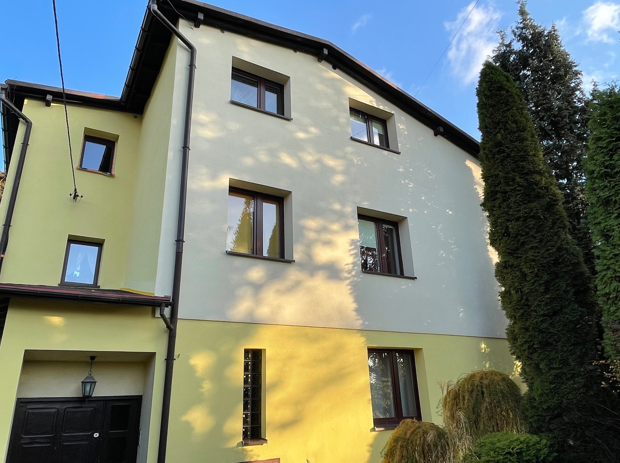 Dwukondygnacyjny dom z elewacją w kolorze żółtym i kremowym, z ciemnymi oknami i rynnami, otoczony zielenią, z widocznymi śladami cieni drzew na ścianie.