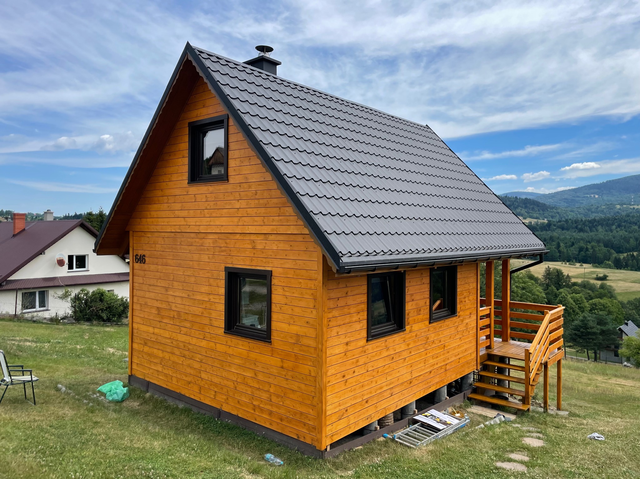 Nowo wybudowany, drewniany dom z szarą, metalową dachówką, numerem '646' i drewnianymi schodami wejściowymi, na tle zielonych wzgórz i błękitnego nieba.