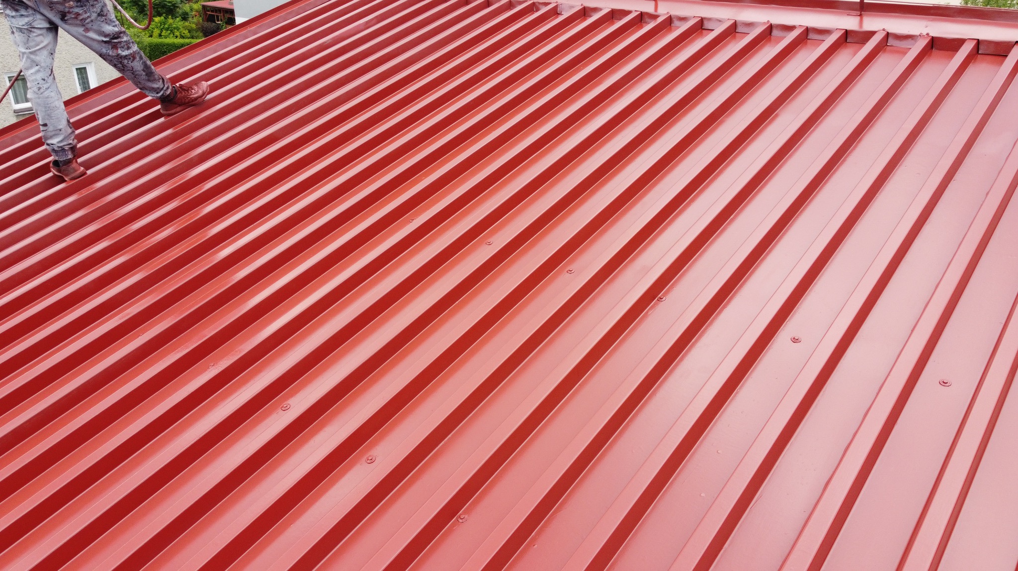 Czerwony dach z blachy trapezowej po renowacji, widoczne nogi pracownika w spodniach pobrudzonych farbą i brązowych butach.