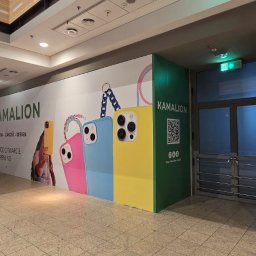 PIXEL WAVE SPÓŁKA Z OGRANICZONĄ ODPOWIEDZIALNOŚCIĄ - Wnętrze galerii handlowej z reklamą sklepu Kamalion, prezentującą kolorowe etui na telefony i informację o otwarciu. Widoczny kod QR i logo sklepu.