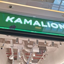 PIXEL WAVE SPÓŁKA Z OGRANICZONĄ ODPOWIEDZIALNOŚCIĄ - Świecący szyld z nazwą 'KAMALION' na zielonym tle, podświetlony zielonymi diodami LED. Widoczne elementy instalacji sufitowej i oświetlenie punktowe.