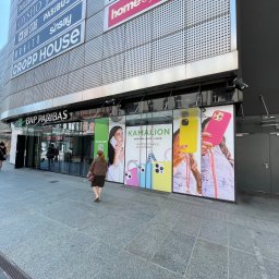 PIXEL WAVE SPÓŁKA Z OGRANICZONĄ ODPOWIEDZIALNOŚCIĄ - Billboard reklamowy sklepu z akcesoriami do telefonów 'Kamalion' na fasadzie budynku w centrum miasta. Widoczni przechodnie.