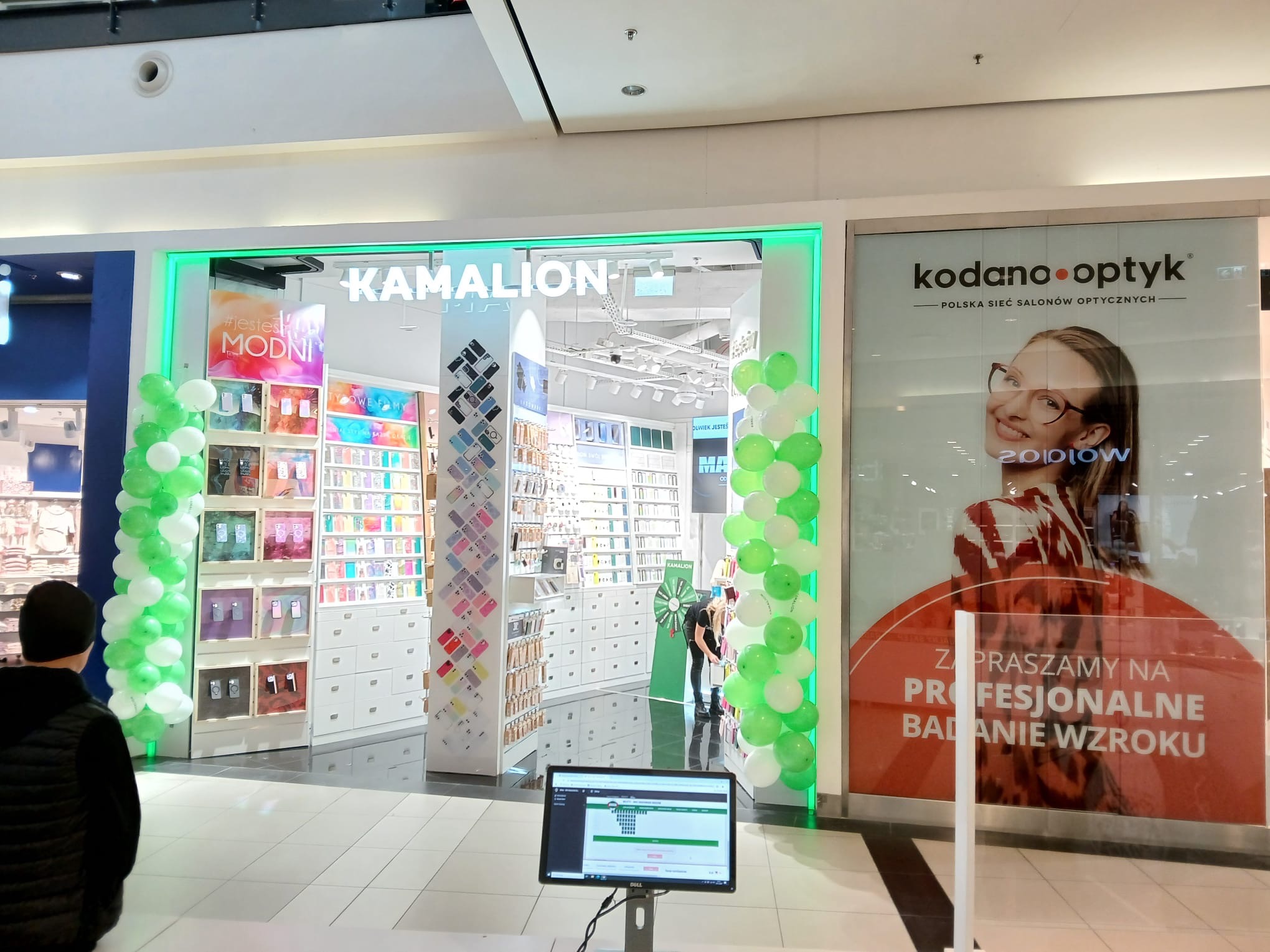 Witryna sklepu Kamalion w centrum handlowym, z zielonym neonowym obramowaniem i dekoracjami z białych i zielonych balonów, obok salon optyczny Kodano Optyk z reklamą badań wzroku.
