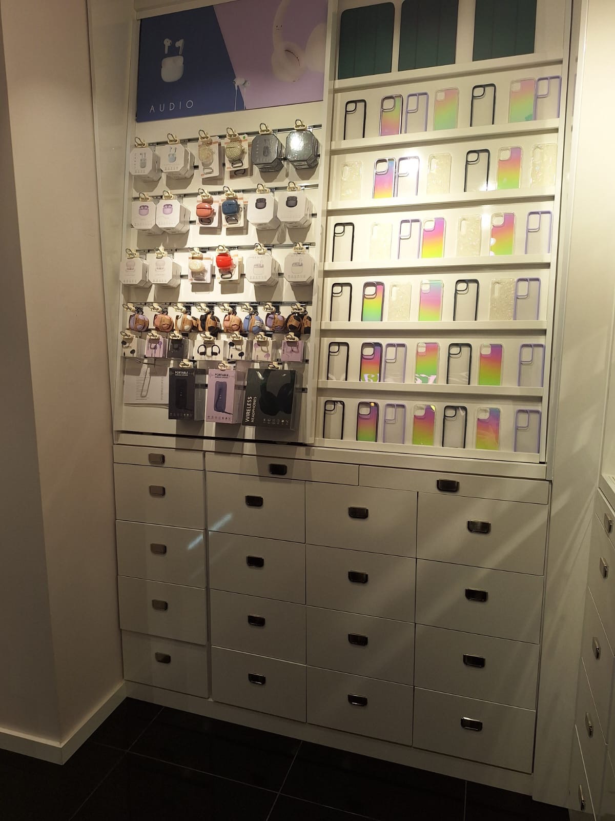 Ekspozycja akcesoriów GSM: etui na telefony z tęczowym gradientem i opakowania słuchawek bezprzewodowych na białym regale z szufladami w sklepie.